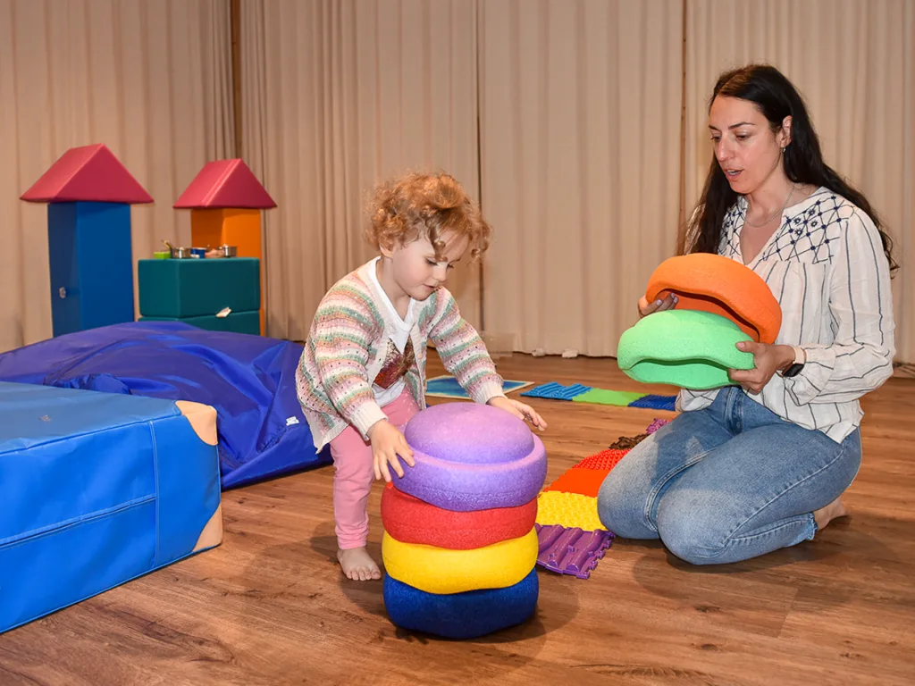 Foto eines Kindes, das sich zusammen mit einer Betreuerin in einem Spielzimmer mit bunten Spielmaterialien befindet